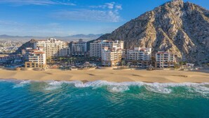 Un hotel costero con varias edificaciones, una playa de arena y una montaña rocosa al fondo.