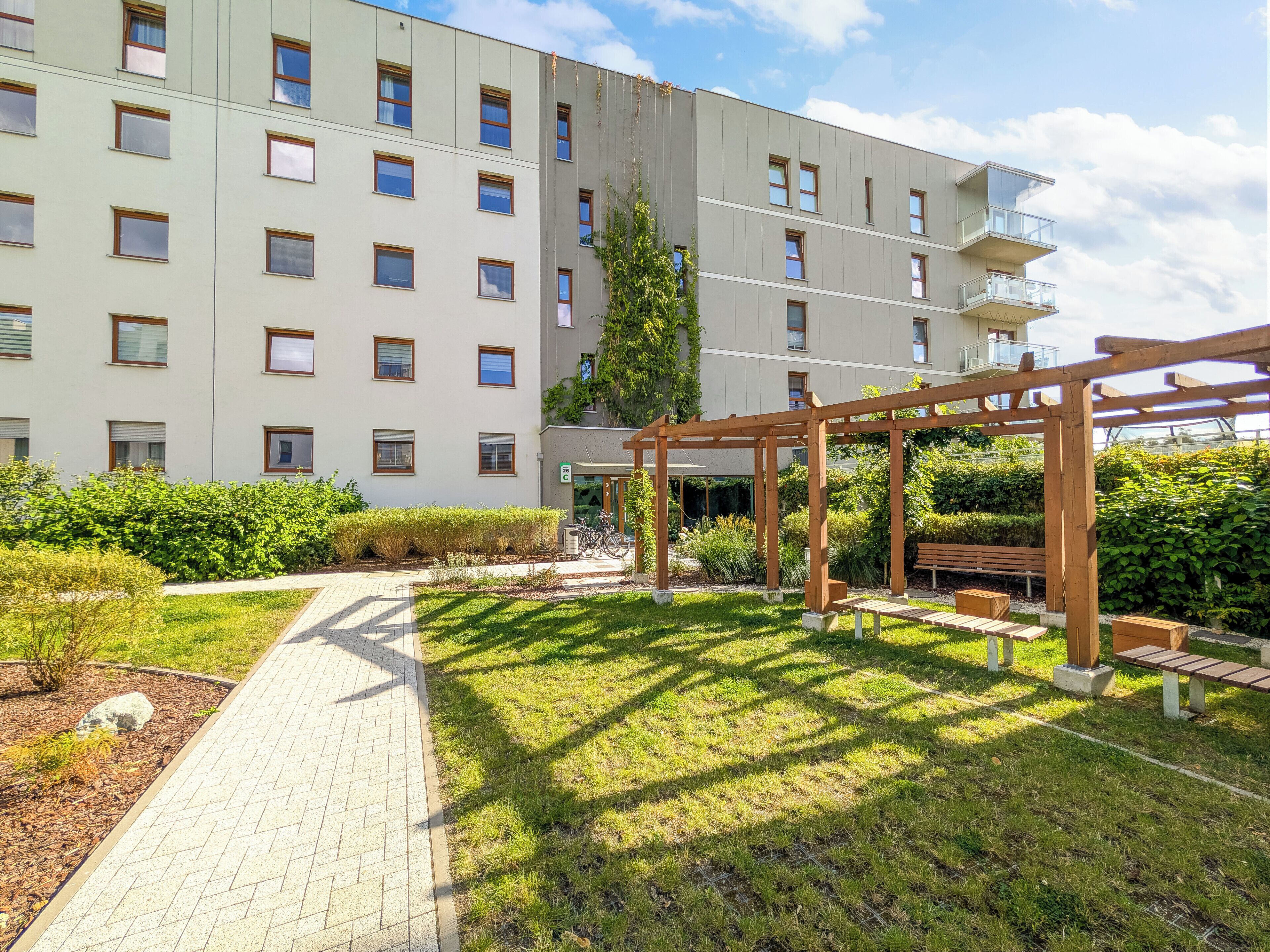 A multi-story building with a garden area featuring a wooden pergola, benches, and a paved walkway.