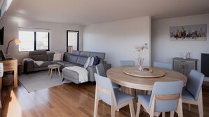 Un salon moderne avec un canapé d’angle gris, une table de salle à manger ronde en bois et des chaises bleues, ainsi qu’un tableau représentant un paysage urbain accroché au mur.