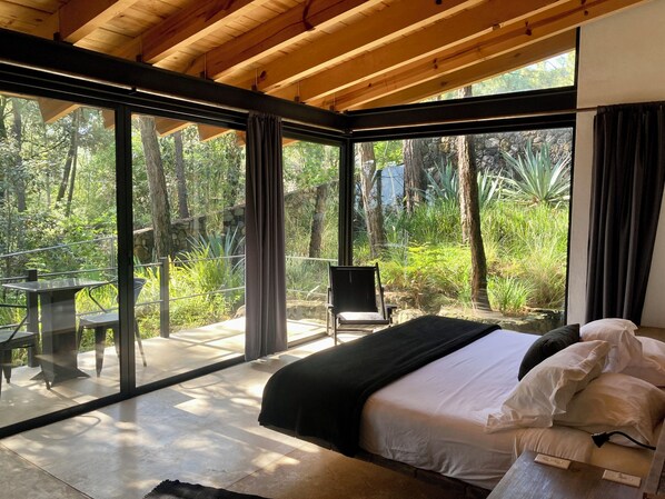 A bedroom with a bed, a chair, and a table, large windows with curtains, and a view of greenery outside.