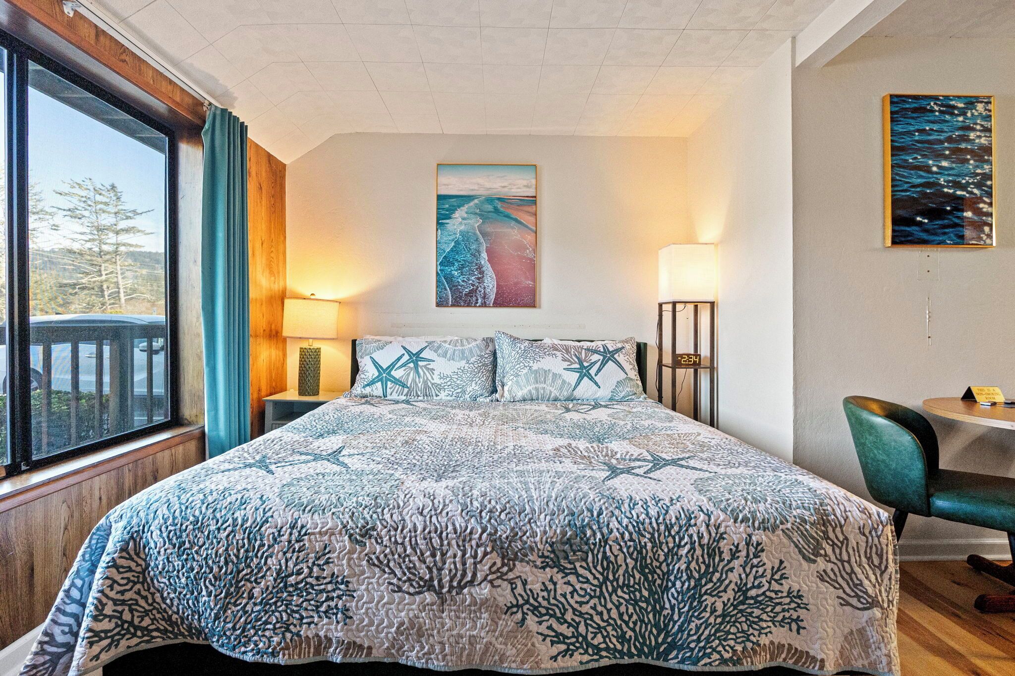 Un dormitorio con una cama grande, una ventana con cortinas, un cuadro en la pared y una pequeña mesa con una silla.