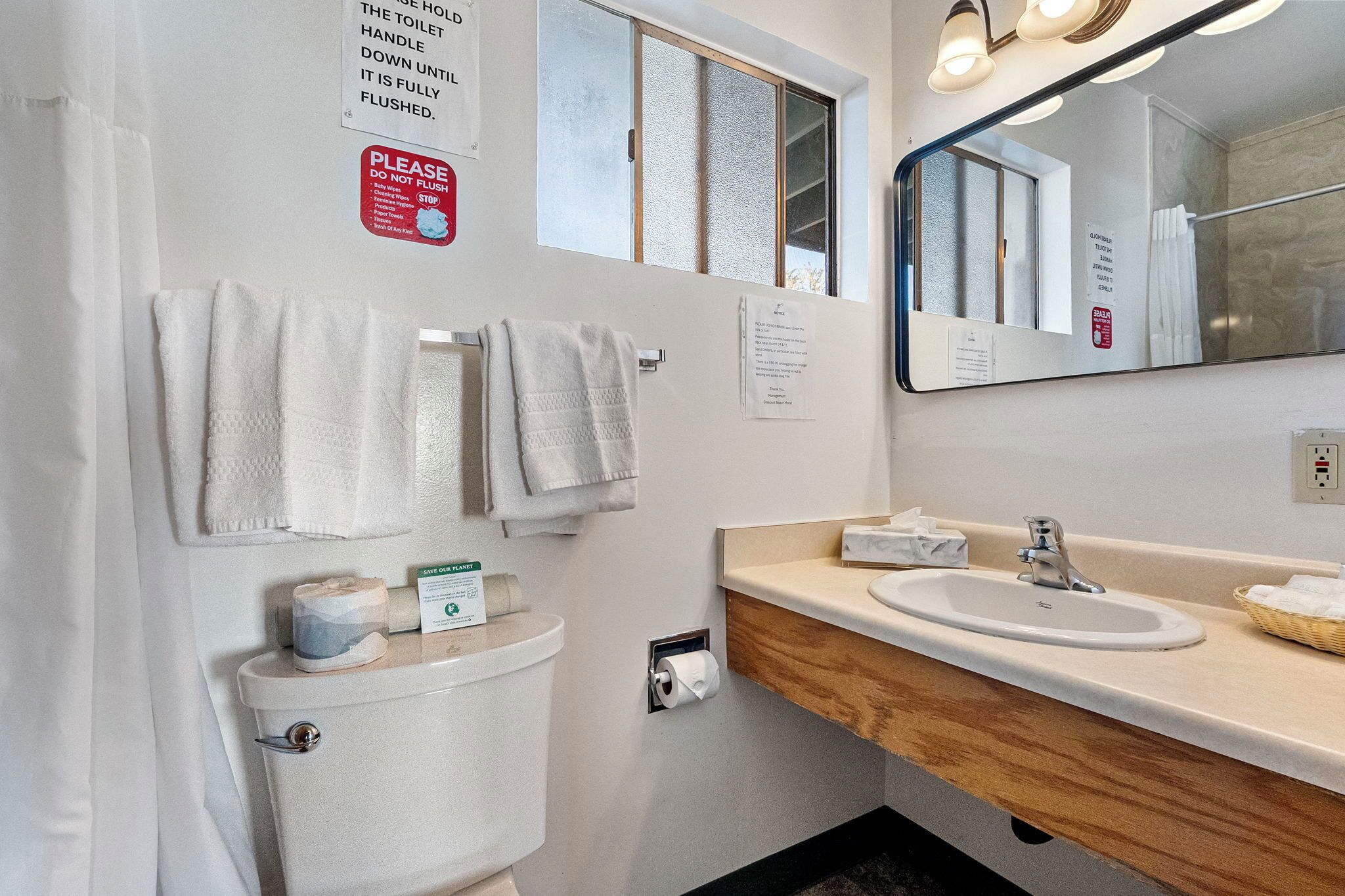 A bathroom with a toilet, sink, and mirror. There are towels hanging on the wall, a window, and a sign about not flushing the toilet.
