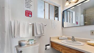 A bathroom with a toilet, sink, and mirror. There are towels hanging on the wall, a window, and a sign about not flushing the toilet.