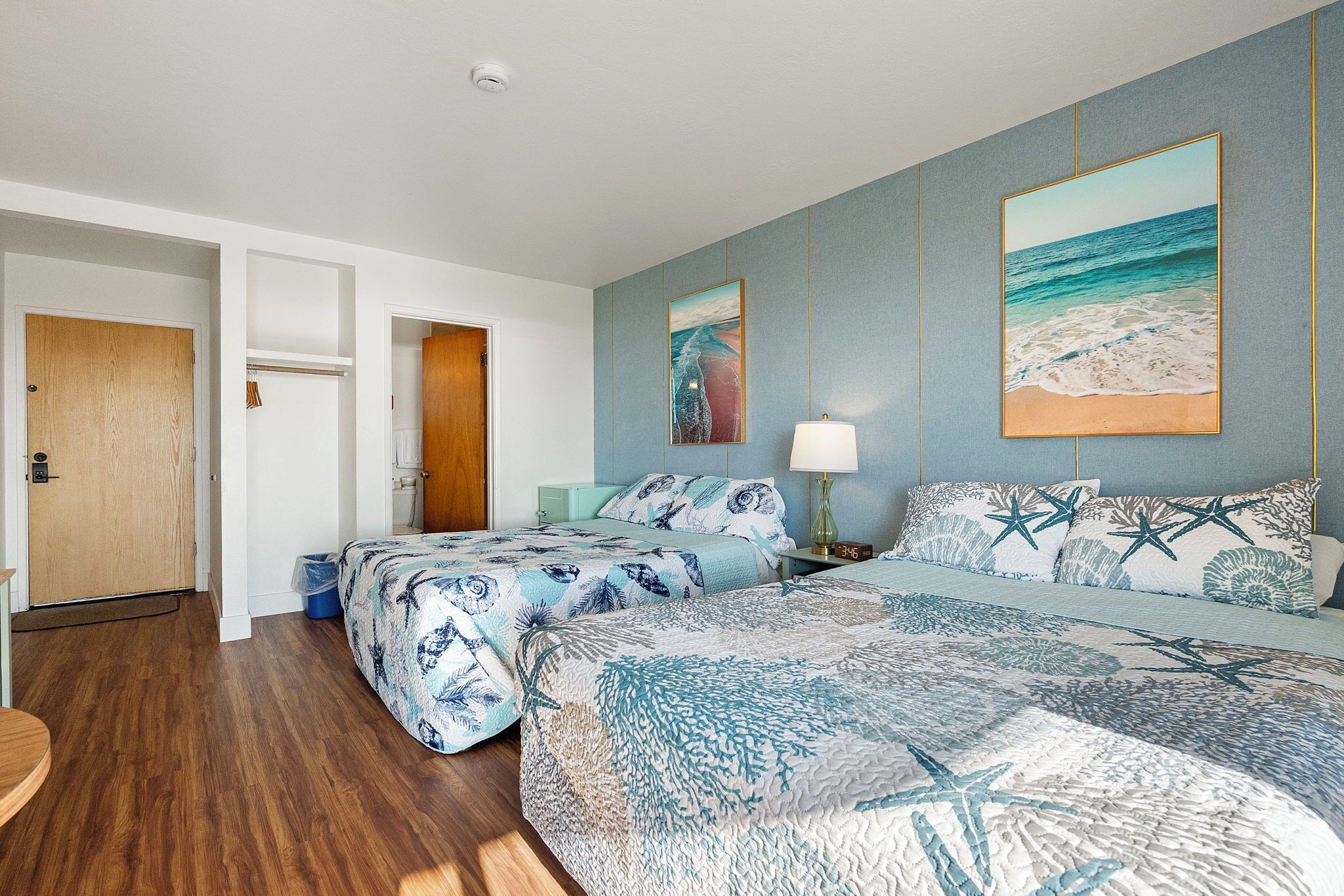 Een hotelkamer met een groot bed, twee kleinere bedden, een schilderij van de oceaan en een houten deur.