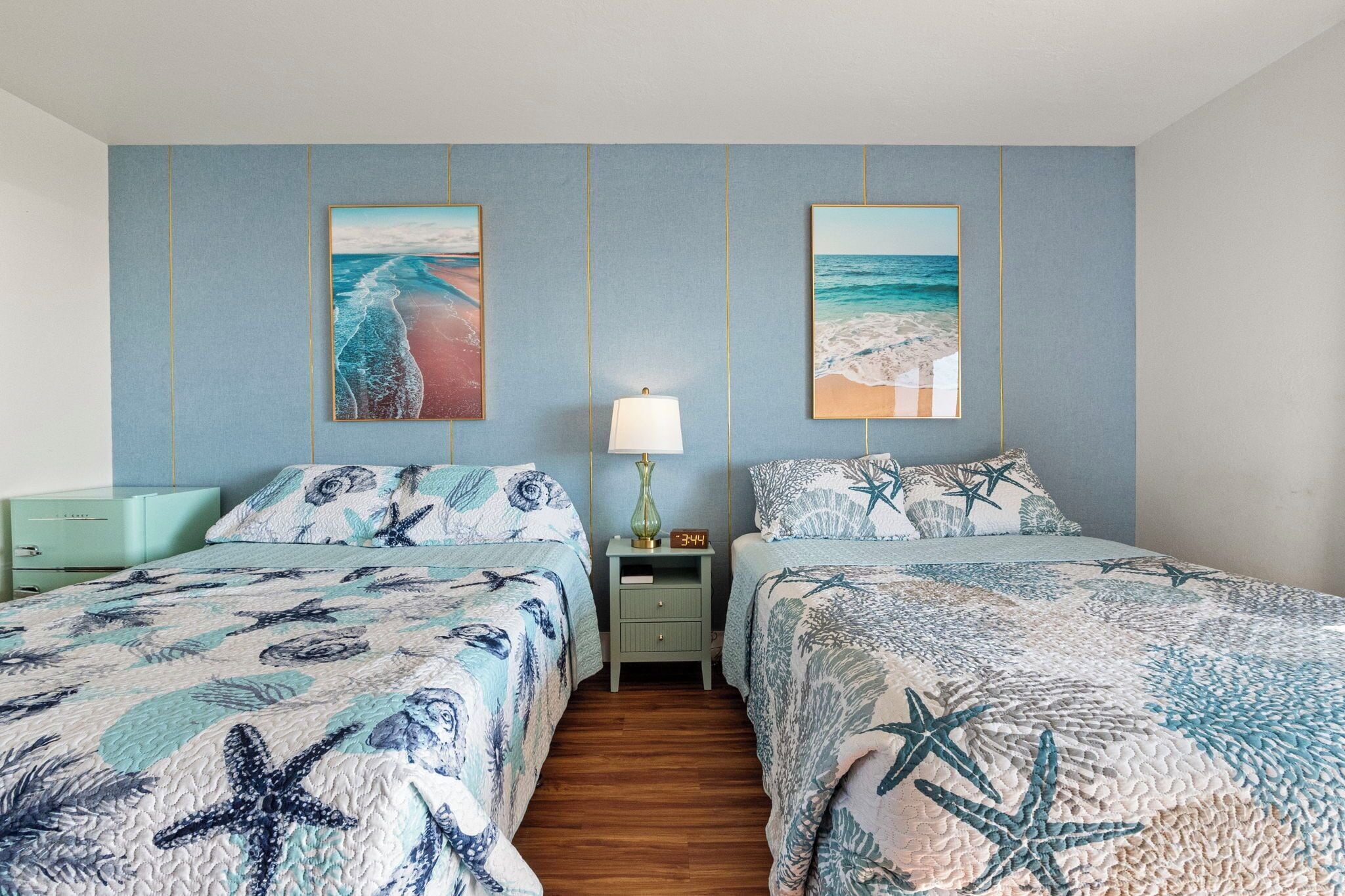 A bedroom with two beds, each with a starfish-themed bedspread, a nightstand with a lamp, and two framed pictures of beaches on the wall.