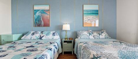 A bedroom with two beds, each with a starfish-themed bedspread, a nightstand with a lamp, and two framed pictures of beaches on the wall.