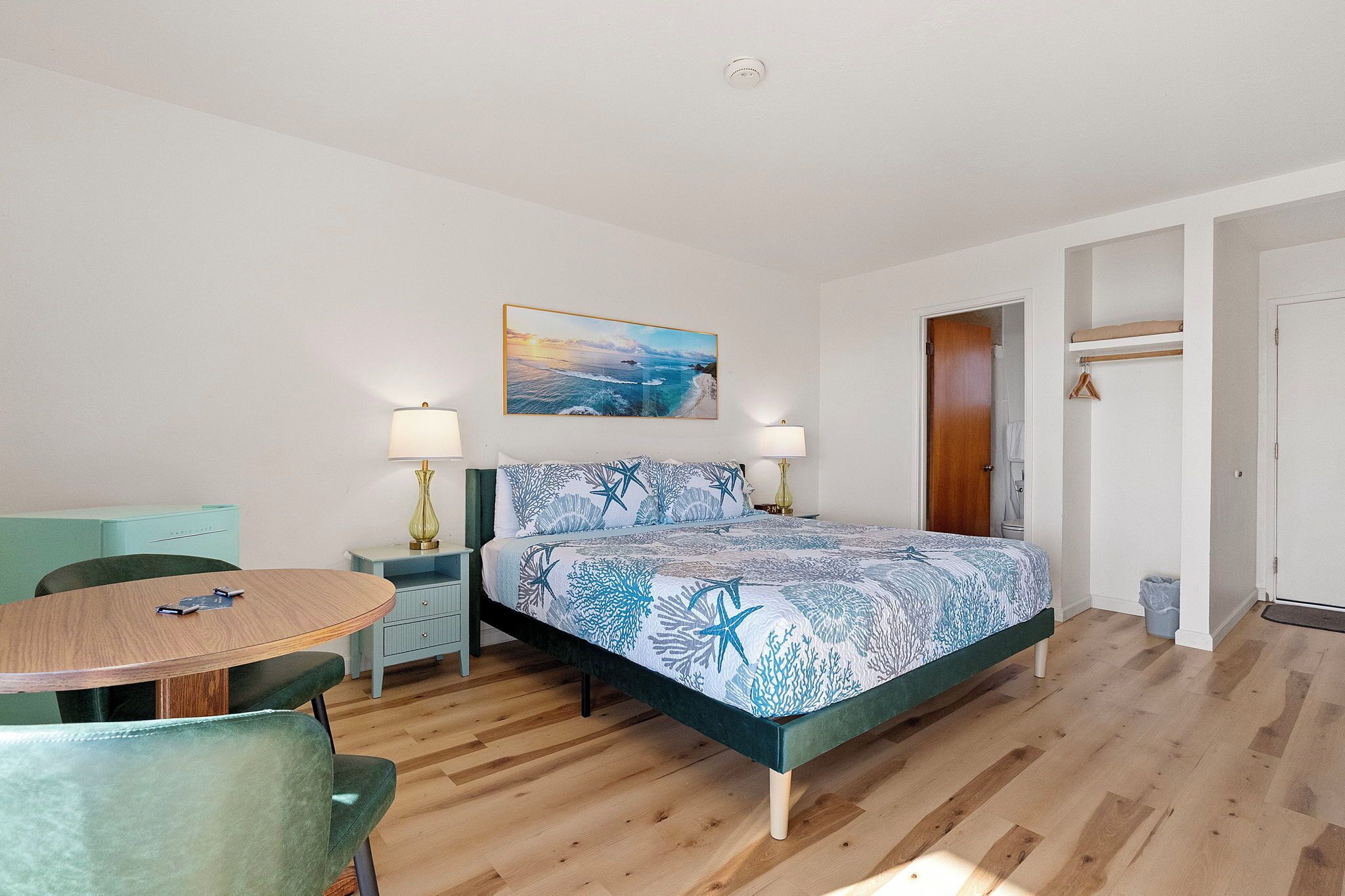A bedroom with a bed, a desk, and a chair. There is a painting on the wall and a door to the right.