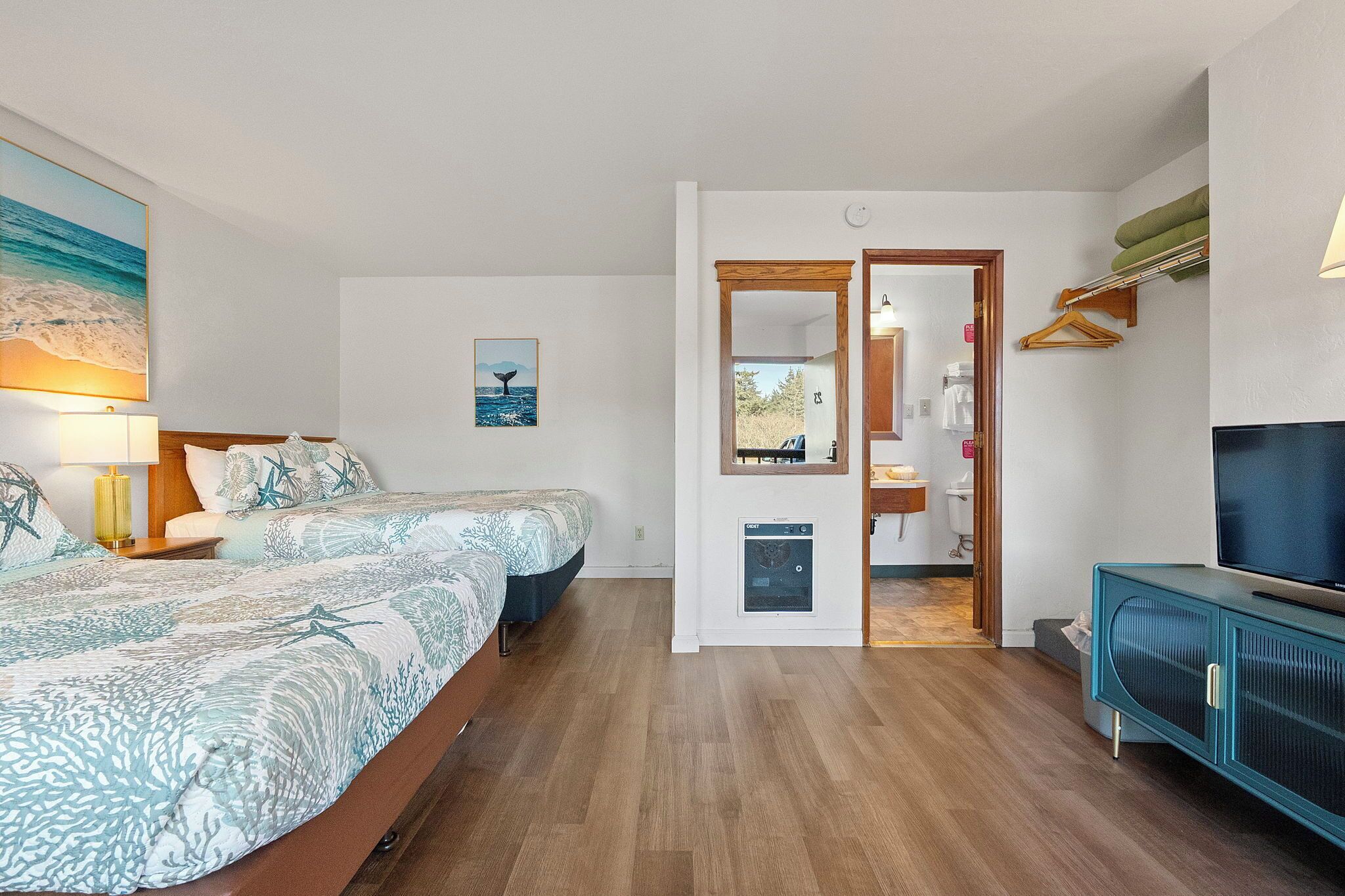 A bedroom with a bed, bedside table, lamp, and artwork. There is a hallway leading to a bathroom and a living area with a TV and cabinet.