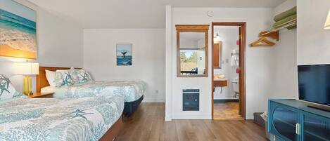 A bedroom with a bed, bedside table, lamp, and artwork. There is a hallway leading to a bathroom and a living area with a TV and cabinet.