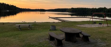 A lakeside park with picnic tables, a swing set, and a dock extending into the water.