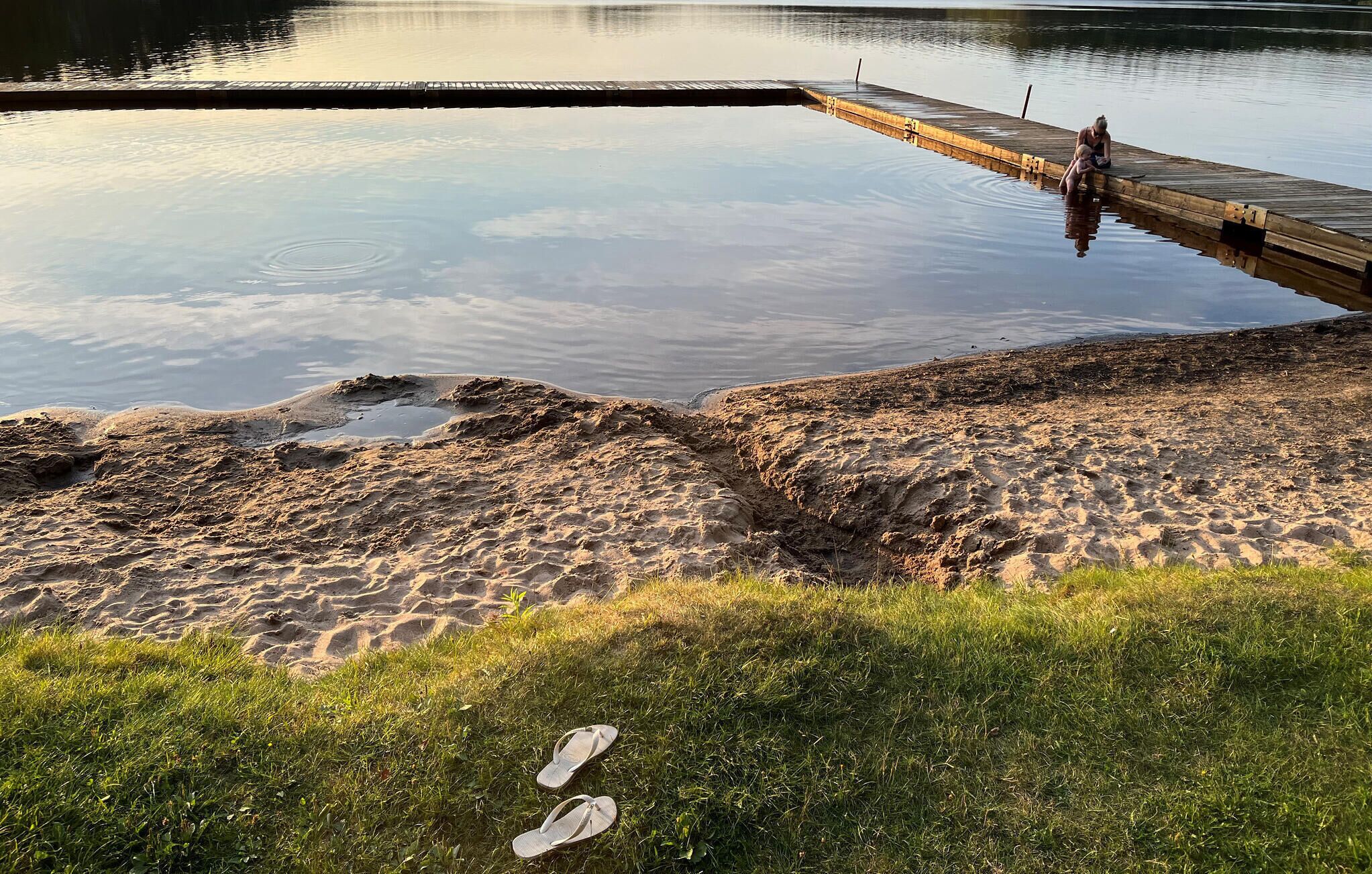 A sandy beach with a wooden dock extending into calm water, and a pair of flip-flops on the grass.