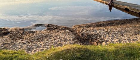 A sandy beach with a wooden dock extending into calm water, and a pair of flip-flops on the grass.