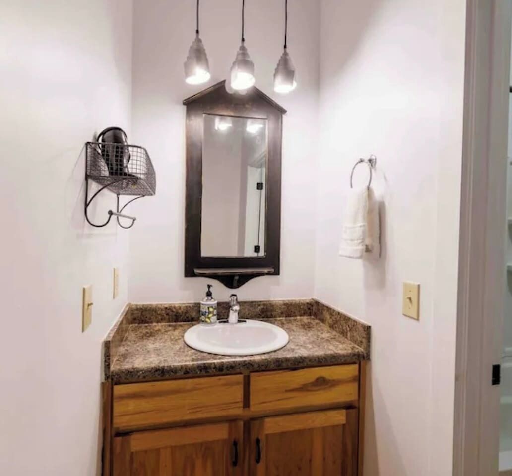 Un baño con encimera de granito, un lavamanos blanco, armarios de madera, un espejo montado en la pared y luces colgantes.