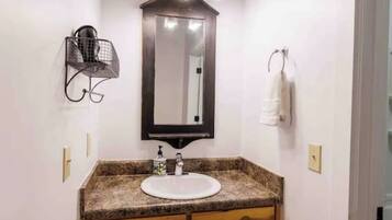 Un baño con encimera de granito, un lavamanos blanco, armarios de madera, un espejo montado en la pared y luces colgantes.