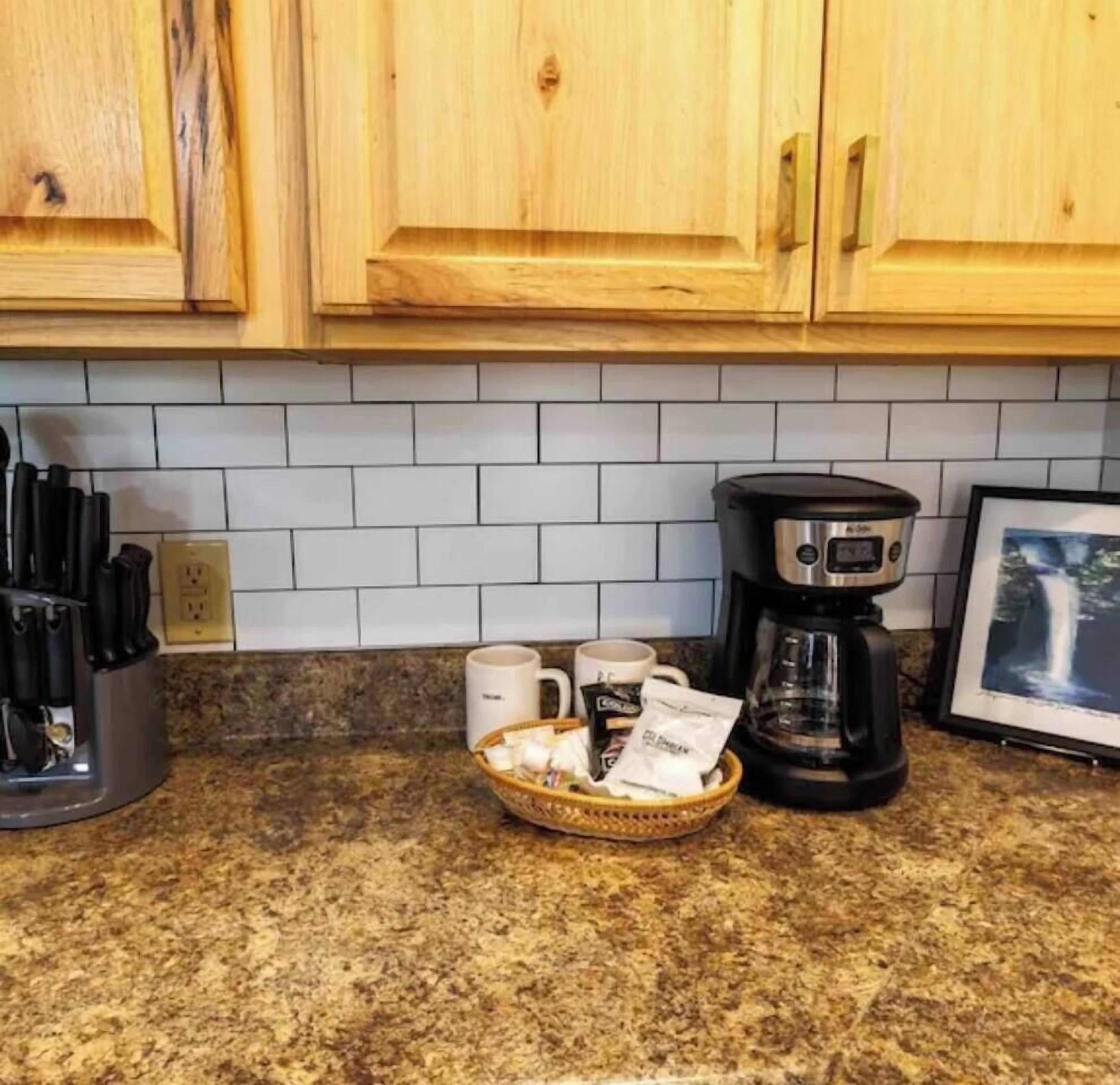 A kitchen with wooden cabinets, a granite countertop, a coffee maker, mugs, and a basket with food items.