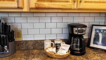 A kitchen with wooden cabinets, a granite countertop, a coffee maker, mugs, and a basket with food items.