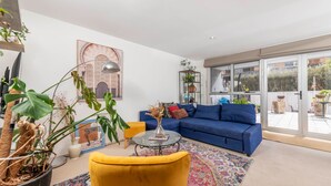 Un salon moderne avec un canapé bleu, un fauteuil jaune et une table basse en verre. On y trouve des plantes en pot et une grande fenêtre donnant sur l’extérieur.