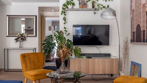 Un salon avec un téléviseur, un fauteuil jaune, une table basse en verre et une plante en pot.