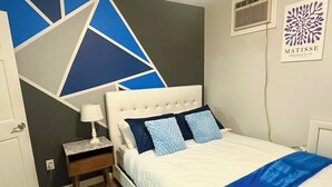 Ein Schlafzimmer mit einem weißen Bett, blauen Kissen, einem Streifen Teppich und einem geometrischen Wandmuster.