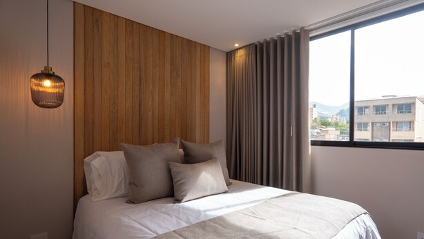A modern bedroom with a wooden panel wall, a large bed with white bedding, and a view of buildings through a window with curtains. - Elegant 3 bedroom apartment in quiet and central area 201 (Medellín)