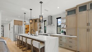 A modern kitchen with a central island, bar stools, wooden cabinets, and stainless steel appliances. - Brand New w/ Luxury Amenities | AC | Fire Pit | Walk to Beach, Bay, Shops, Eateries (San Diego)