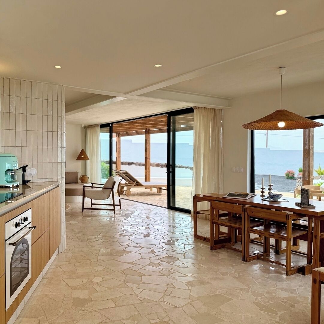 A modern kitchen with a coffee machine, a dining area with a table and chairs, and a view of the beach through large sliding doors.