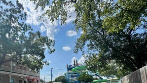 Un parking avec un parc aquatique en arrière-plan, un bâtiment portant une enseigne indiquant « Pizza maison chez D », et un autobus au loin.