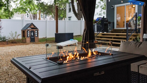 A modern outdoor seating area with a built-in fire feature, a covered patio, and a view of a house in the background.