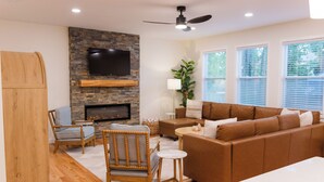 A living room with a stone fireplace, a flat-screen TV mounted above it, a brown leather sofa, a wooden chair, and a small round table.