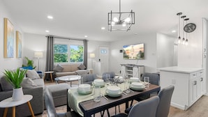 A modern, well-lit living room with a dining area, grey sofas, a coffee table, and a TV displaying the Capitol building. - Upscale Old Town Views 3BD Lux (Alexandria)