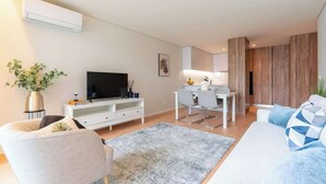 A modern living room with a flat-screen TV, a dining area with a white table and chairs, and a sofa with patterned cushions.