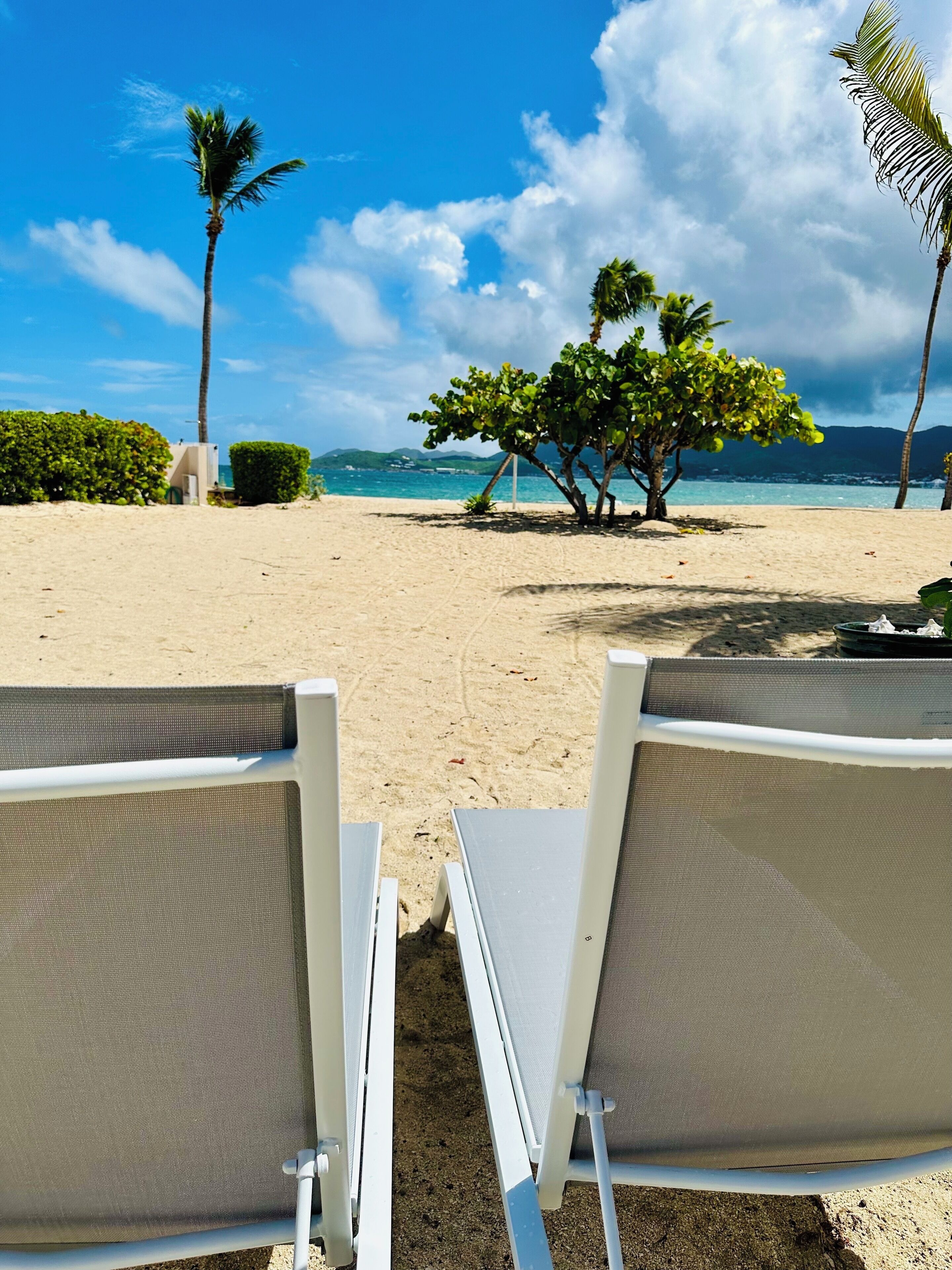A beach with two white lounge chairs facing the ocean, palm trees, and a clear sky.