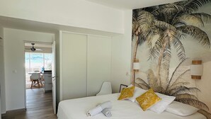 A bedroom with a bed, a headboard with a palm tree mural, a wooden floor, and a view of the outside. - Salty Beach - NBBC- 1 bedroom beachfront (Marigot)