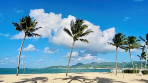 A beach with palm trees, a clear sky, and a distant view of mountains and a city. - Salty Beach - NBBC- 1 bedroom beachfront (Marigot)