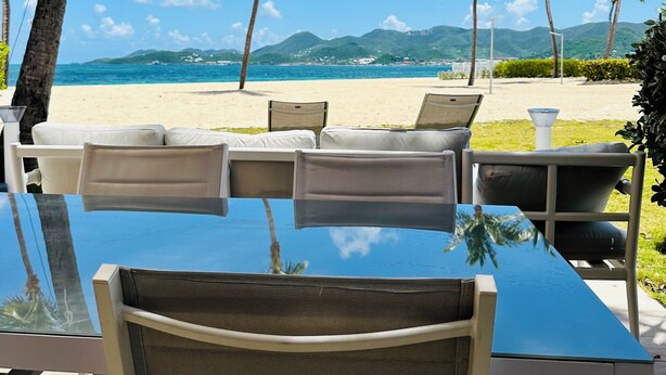 A beachside dining area with a glass table and chairs, overlooking a sandy beach and palm trees. - Salty Beach - NBBC- 1 bedroom beachfront (Marigot)
