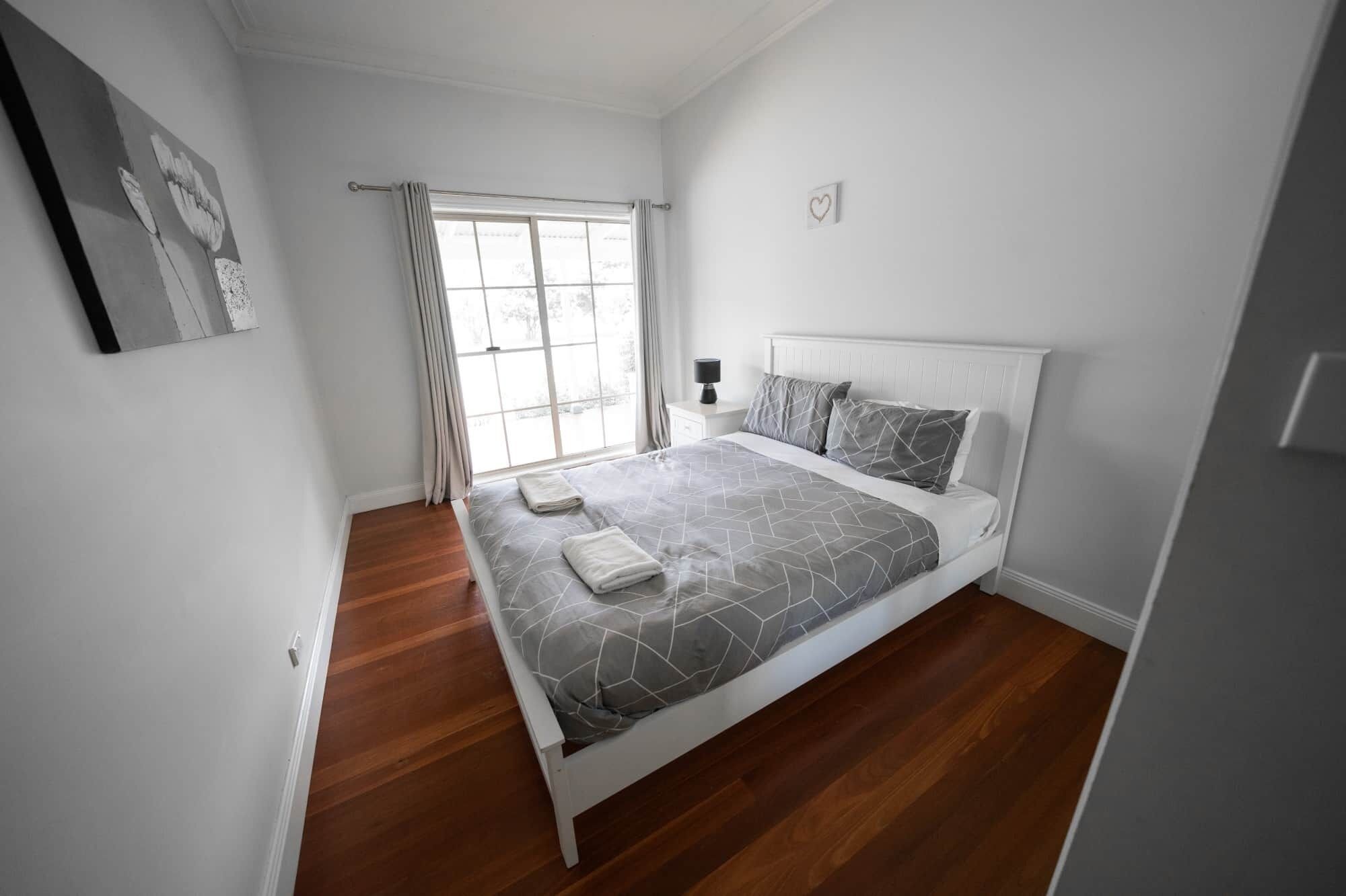 A bedroom with a bed, a nightstand, a window with curtains, and a wall-mounted heart decoration.