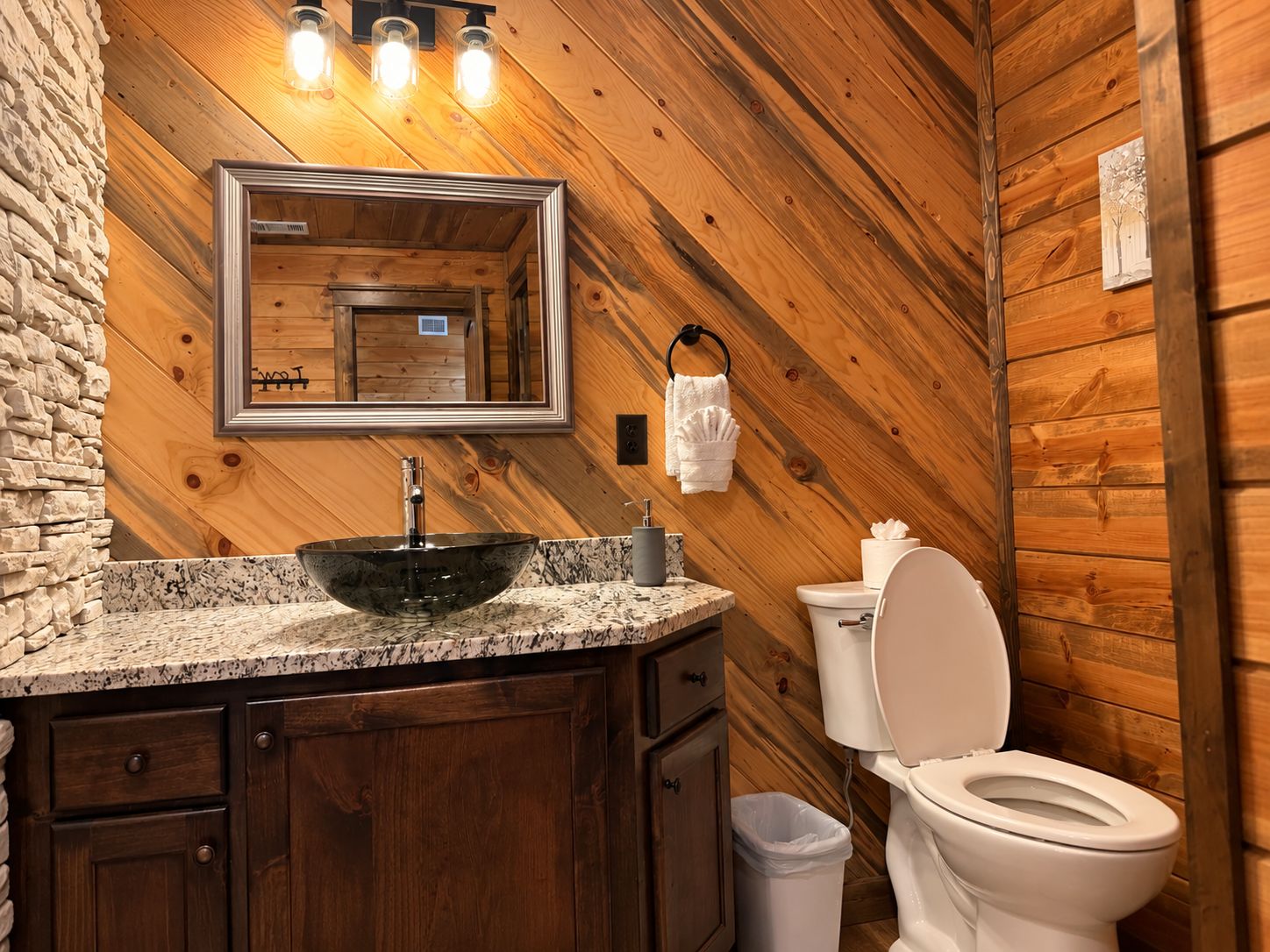 A wooden cabin-style bathroom with a granite countertop, a vessel sink, a toilet, and a mirror with a towel ring.