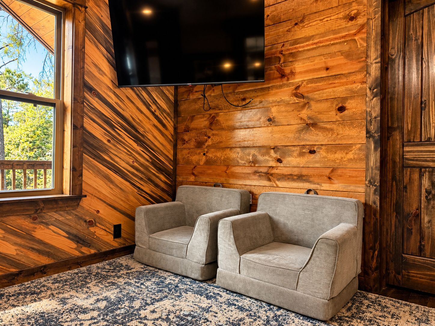 A room with a large flat-screen TV mounted on a wooden wall, two grey armchairs, and a patterned rug.
