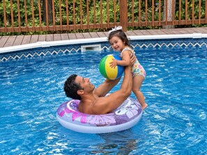 Un hombre y un niño en una piscina con una cubierta de madera y follaje al fondo.