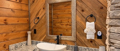 A bathroom with a wooden wall, a large mirror, a vessel sink, a towel ring, and a towel.