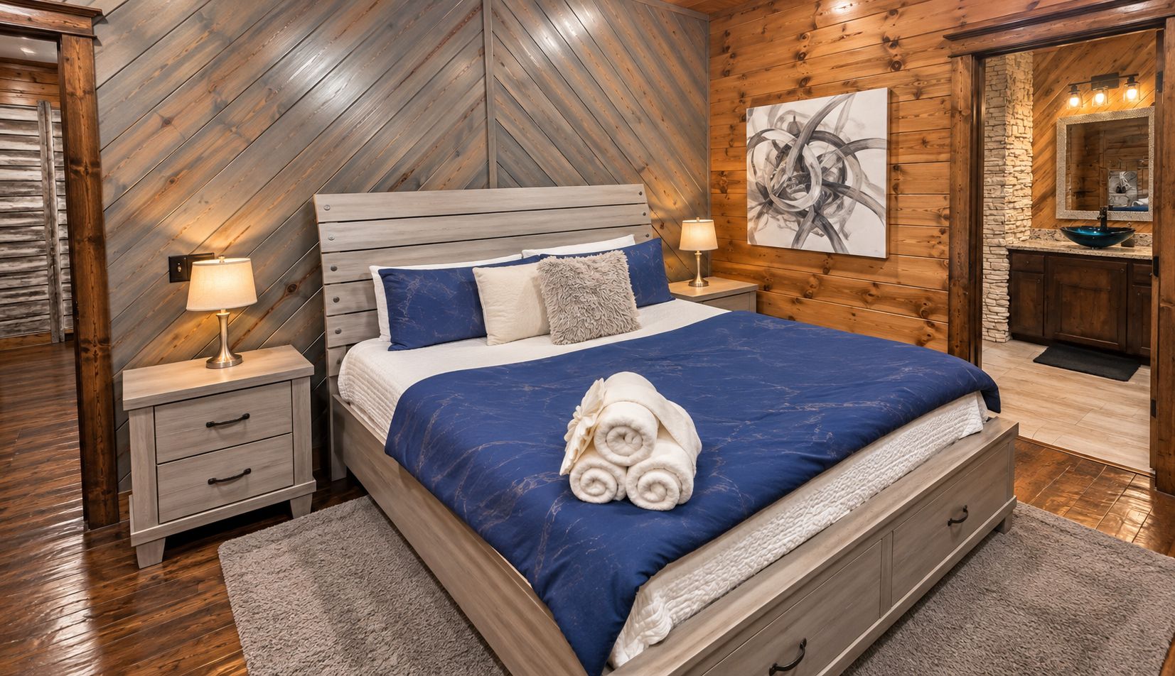 Un dormitorio con una cama, dos mesitas de noche, una mesita auxiliar, un cuadro en la pared, y un baño con lavamanos y espejo.
