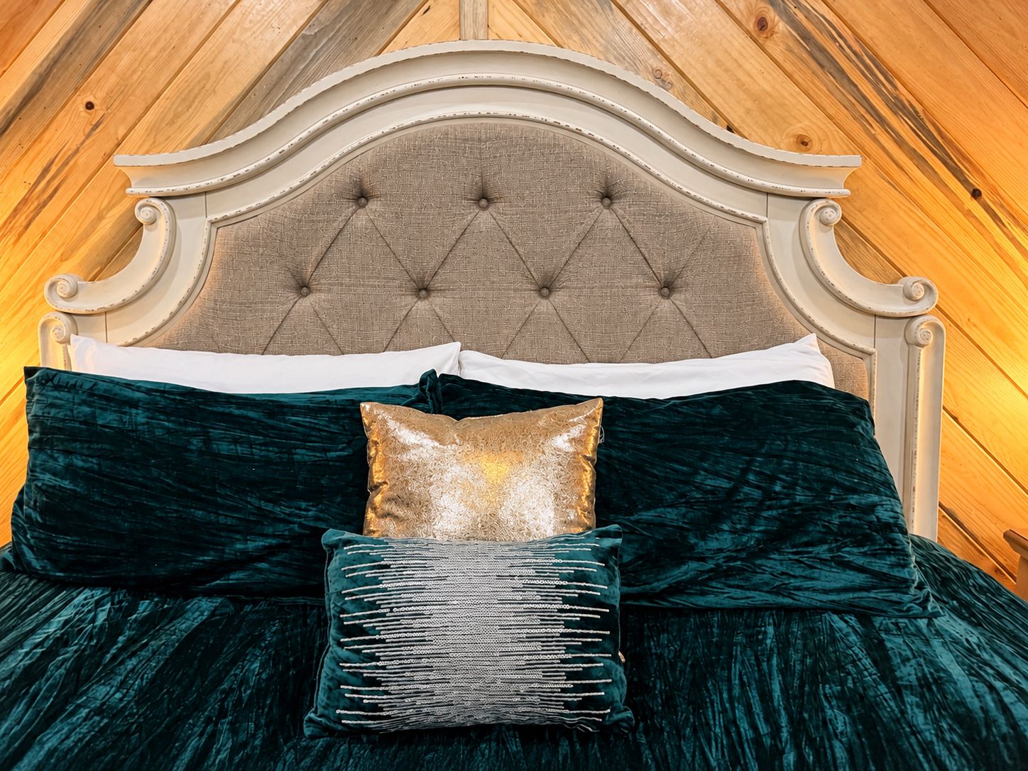 Una cama con cabecera capitonada, almohadas verde oscuro y doradas, y una almohada decorativa tejida.