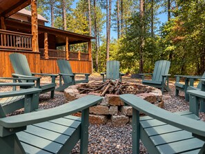 En stuga av trä med en veranda och Adirondack-stolar placerade runt en eldstad.