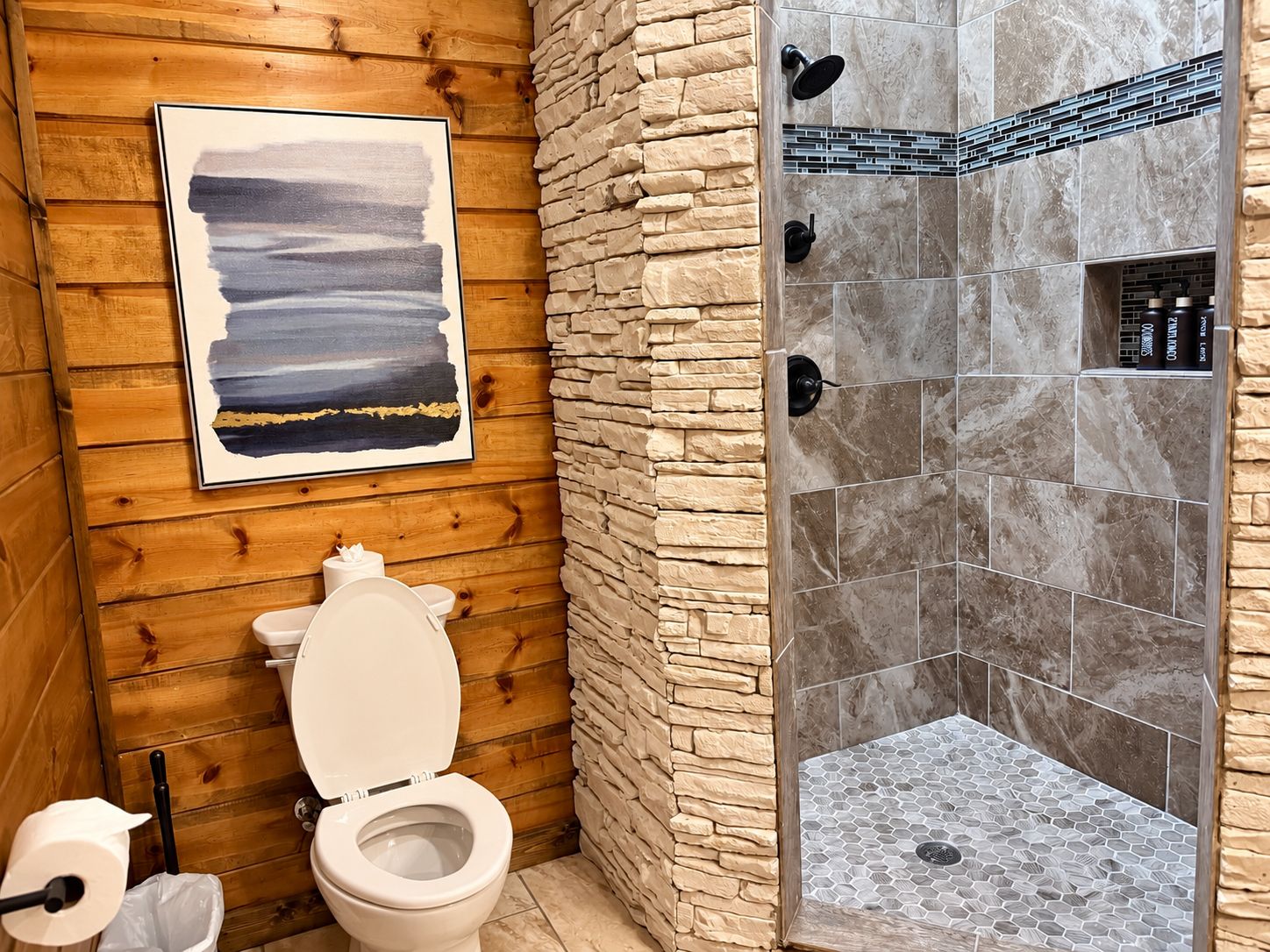 A bathroom with a wooden wall, a white toilet, a shower with stone tiles, and a framed picture on the wall.