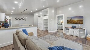 Ein modernes Wohnzimmer mit Sofa, Couchtisch und Fernseher auf einem Lowboard. Es gibt eine Küchenzeile mit Arbeitsplatte und Schränken.
