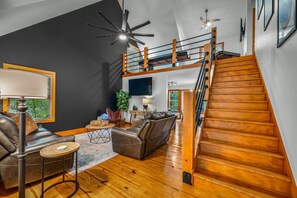 Amplia sala de estar con escalera de madera, muebles de cuero y ventilador de techo.