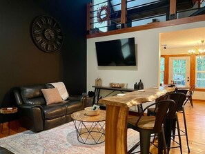Una sala de estar moderna con un sofá de cuero negro, una mesa de comedor de madera con sillas, un reloj de pared grande y un televisor de pantalla plana montado sobre la mesa.