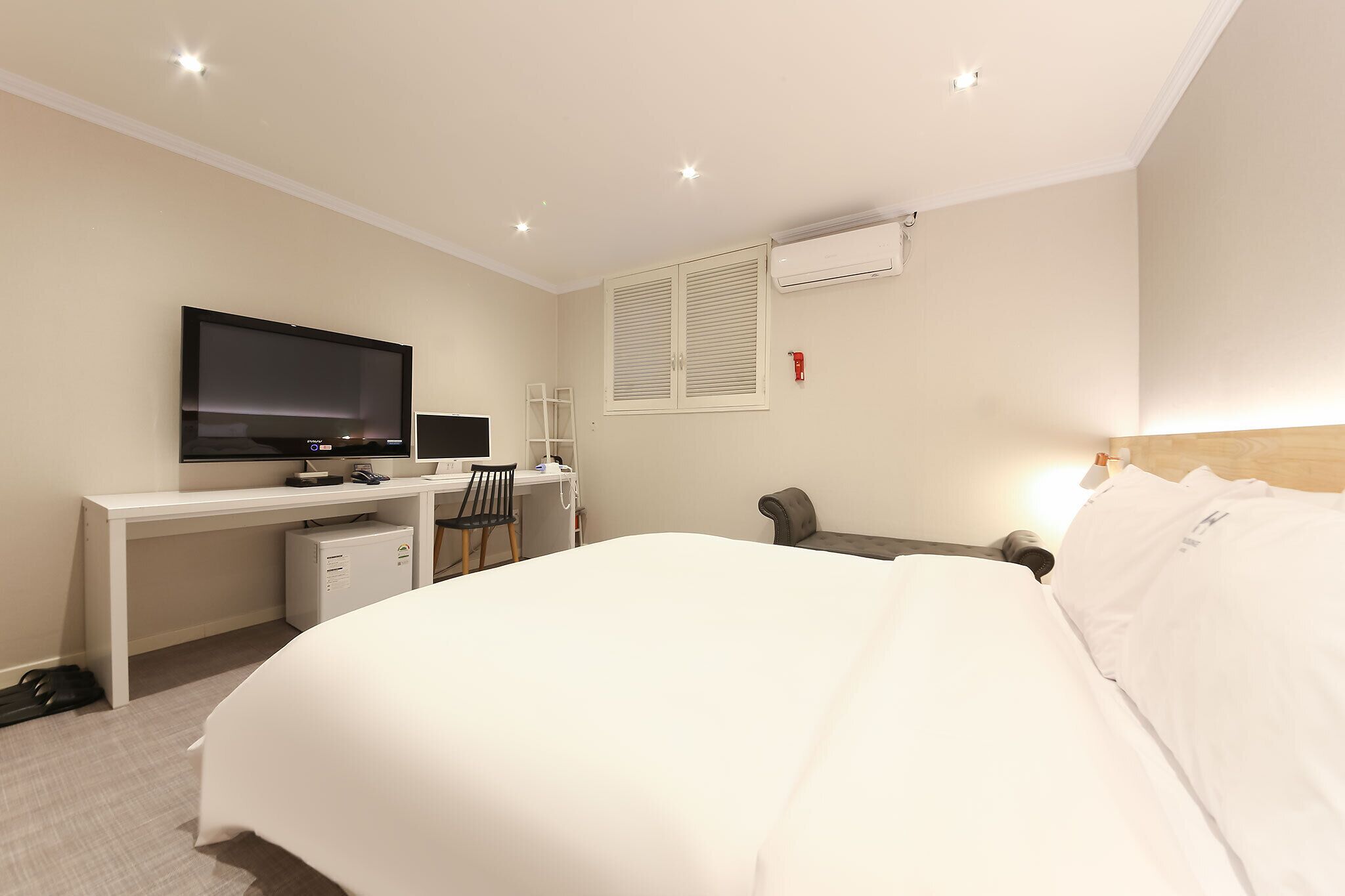 A hotel room with a bed, a desk with a computer, a TV, and an air conditioning unit.