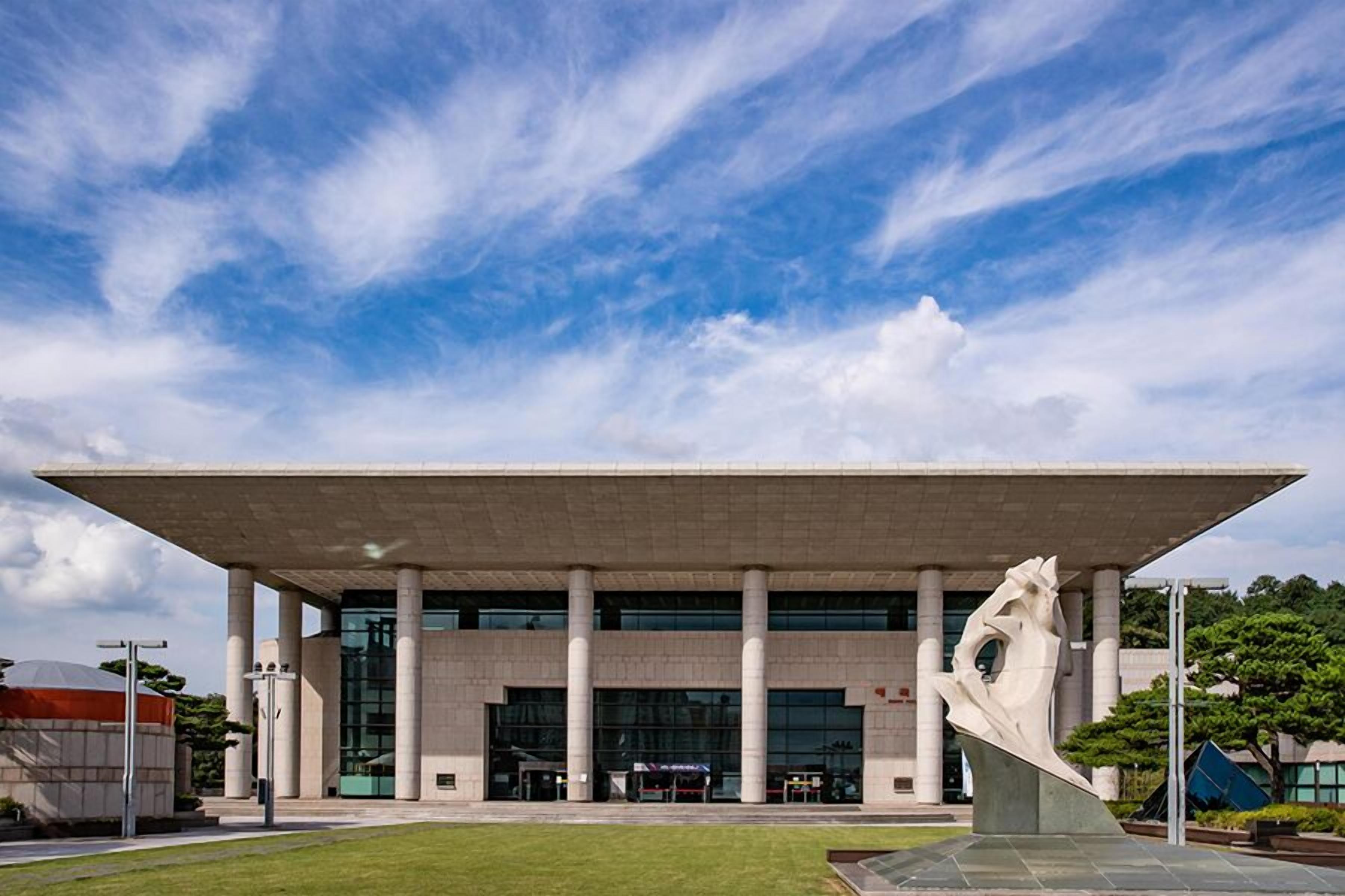 A modern building with a flat roof, large columns, and a central sculpture.