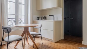 Une cuisine de taille réduite avec des meubles blancs, une table ronde en bois, deux chaises grises et une fenêtre donnant sur l’extérieur.